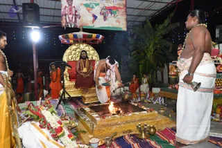 Night ritual at an open-sided temple hall lit by a bright stage lamp and strings of coloured fairy lights. In the centre, a fire burns in a square stone homa pit set on a striped mat. A bare-chested priest in white dhoti with a pink-and-white cloth over his shoulder bends over the fire, holding a ladle. Beside him, a man in a white dhoti with a bell in hand stands at the right edge, and a shirtless attendant with a garland stands at the left. Behind the fire, a seated figure in a red robe and ornate gold necklace sits on a gilded throne with a red cushion and an ornate yellow canopy above. Brass pots, flower garlands and offerings fill the foreground. Several orange-clad and white-clad attendants stand in the background.