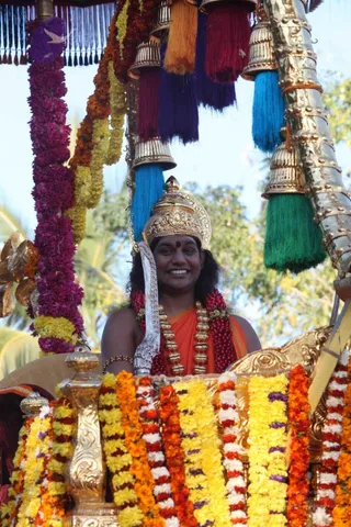 A smiling man rides in an ornate ceremonial chariot or palanquin decorated with layered marigold and mixed flower garlands in orange, yellow, magenta and white. He wears a gold crown with a central finial, a deep orange saffron garment, a long gold necklace, multiple rudraksha-style red beaded malas, and a red bindi on his forehead. Above him hang silver bells and tassels in blue, green, purple and red from a silver-and-gold canopy pole. A silver ceremonial sword or staff rests beside him. The setting is outdoors with trees and sky behind. Swamiji (probable) is shown in the chariot with saffron clothing, crown and rudraksha malas, and the caption identifies the photo as Nithyananda Paramashivam.