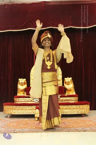 A standing man raises both arms high in a blessing gesture before a dark red velvet drape and a gilded throne flanked by two golden lion figures on red cushions. He wears a rust-brown turban with a gold ornament, gold necklaces and bangles, and a brown and gold garment with a cream shawl draped over one shoulder. A patterned orange and red carpet covers the floor. Swamiji (probable): the figure wears a turban, ceremonial gold necklaces and ornate saffron-brown clothing, consistent with the page's Swamiji reference, and is identified by appearance only.