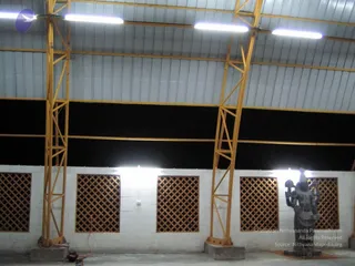 Interior view of a large building under construction, with a corrugated metal roof supported by yellow steel trusses and columns. Fluorescent tube lights hang from the frame. Along the white block wall are latticed brick windows, and a dark grey stone deity murti stands on the right near the wall. No people are visible; the floor is bare concrete with a coiled orange cable at lower left.