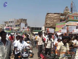 A dense street procession in daytime moves through a busy Tamil Nadu town road lined with traffic. Many men in white shirts and dhotis and casual shirts walk and ride motorbikes, with some women in saris at right. Several large posters and banners on the right show a figure wearing an orange-red turban and saffron-toned clothing, with a red-and-yellow poster reading Kovilpatti. A colourful gopuram-style temple tower with painted deities stands at upper right, and a thatched hut and multi-storey buildings line the street. Buses and trucks crowd the road at left.