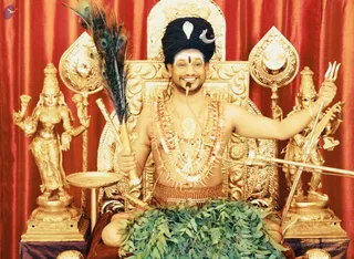 A seated man on a gold ornate throne with red draped curtains behind, smiling broadly with a microphone near his mouth. He wears a tall black turban with a white ornament and crescent, a white-and-black forehead marking, layered red and white beaded malas and a large ceremonial pendant, and a beaded waist cord; his upper body is bare. He holds a peacock-feather fan in one hand and a trident in the other, with green leaves draped across his lap. Flanking him are gold statues of deity forms, one holding a lotus-like object and another with a veena-like instrument, and a gold oil lamp stands at left.