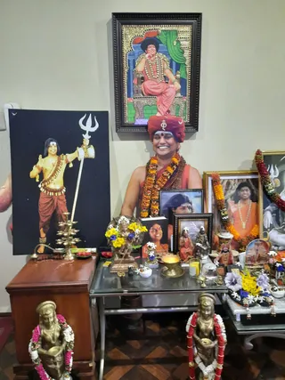 Indoor shrine arrangement against a plain cream wall. A standing woman in a red-orange turban and saffron-orange robes, wearing orange-and-yellow marigold garlands and rudraksha malas, smiles at the centre of the shrine. Above her hangs a framed tanjore-style painting of a seated robed figure in pink-orange clothing on a gold throne. To the left stands a large poster of a standing figure with a trident and orange dhoti. Brass bronze statues of deities with pink and white garlands stand on the floor in front, and a brass oil lamp, flower arrangements, framed portraits and a bronze deity figure sit on a glass table.