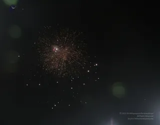 A night photograph of a burst of orange and white fireworks against a dark sky. Sparks radiate outward from a bright central point in a dense spherical spread, with scattered small lights trailing below. No people, figures or structures are visible. The bokeh of out-of-focus lights appears at the edges.