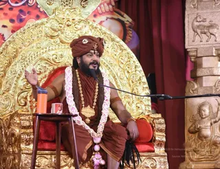 A seated man gestures with his right hand while speaking into a microphone on a stand. He sits on an ornate gold throne with red cushions, wearing a rust-brown turban with a small emblem, brown-orange robes, a rudraksha mala with a large gold pendant, and a pink-and-white flower garland. A small wooden side table holds an orange flask and a cup. Behind him are red curtains and a carved golden pillar with a figure. The person matches the Swamiji (probable) appearance cues of a turban, saffron-toned clothing, rudraksha and gold ornaments, and the page indicates he is at this event.