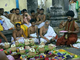 A temple ritual on a stone floor beside a pillar marked A21. Several bare-chested men in white and yellow dhotis sit in rows, some wearing rudraksha malas. In the foreground are coconuts crowned with white flower garlands, set on decorated, colourful bases with mango leaves, alongside brass vessels and pink petals. On the right, a man in an orange-red robe and dark turban holds a plate of pink petals and wears long bead malas and a shawl. Beside him sits an older man in white with a tilak and a green-bordered cloth. The scene suggests a temple puja or abhishekam preparation.