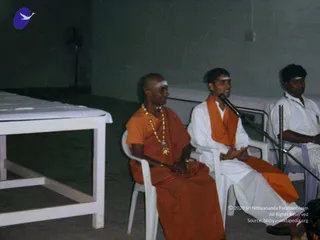 Indoor photo in a dim hall with pale walls. Three men sit on white plastic chairs. The man on the left is shaved-headed, wears an orange robe, a gold-coloured beaded necklace and white tilak marks, and looks toward the centre. The man in the middle wears white clothes with an orange shawl over his shoulder, speaks into a microphone on a stand, and gestures with his hands. The man on the right wears white and sits behind them. A white-covered table stands at left and a fan is visible in the background. No clear identification of the speaker is made from appearance.