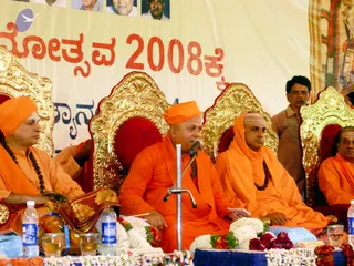 A stage event with several saffron-robed men seated on ornate gold-and-red thrones in front of a large cream banner with Kannada text in red. The central figure in orange robes and an orange turban sits at a microphone stand, speaking, with his hands resting on a white paper. Beside him, a bald man in orange robes and a rudraksha mala sits attentively. On the left, a bearded man in an orange turban and a long rudraksha mala sits beside a draped cloth and a bowl of fruit. Behind them, a man in a brown shirt stands near the throne, and white jasmine garlands and red flowers decorate the stage edge. Bottled water and glasses are on the table.