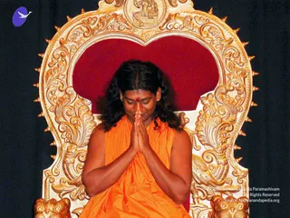 A person in saffron-orange robes sits on an ornate gilded throne with a red velvet back and carved floral scrollwork, with a small figure carved at the crest. The person has long dark hair, eyes lowered, palms pressed together in namaste at the chest, and a red tilak mark on the forehead. The dark backdrop sets off the gold throne and orange cloth. A faint copyright line appears at lower right. Only one physical person is visible.