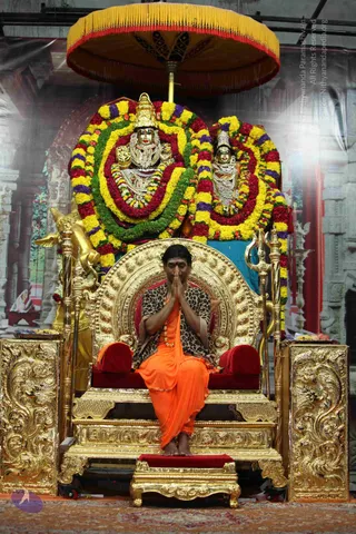 A man seated on an ornate gold throne with red velvet cushions, hands pressed together in namaste with eyes closed. He wears an orange saffron dhoti and a leopard-print shawl over his shoulders, with a beaded necklace. Behind him stand a richly decorated deity murti pair beneath a large yellow-fringed ceremonial parasol, both draped in garlands of yellow, red, green and purple flowers. Golden trident and ornamental pillars flank the throne, and a gold bull figure is visible to the left. The backdrop shows a painted temple-style archway.