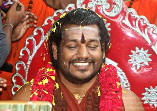Close-up photo of a smiling bearded man with eyes closed, seated in front of an ornate red and silver throne. He wears a red garland of flowers with yellow petals scattered over it, a white forehead marking with a red vertical tilak, a rudraksha mala, a gold necklace and a red-brown shawl. Blurred orange-robed devotees and a hand holding a phone appear at the left edge.