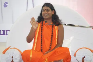 A photograph of a seated man in saffron-orange robes with gold borders, wearing a long rudraksha mala and a red bindi, with long swept-back hair. He sits cross-legged on a white cushioned seat with orange-trimmed bolsters, one hand raised mid-gesture while speaking into a microphone on a flexible stand. A white backdrop and a purple banner with partial lettering are behind him. The appearance is consistent with the a person candidate named on the page, based on hair, saffron clothing and rudraksha mala, so the identity is probable rather than confirmed.