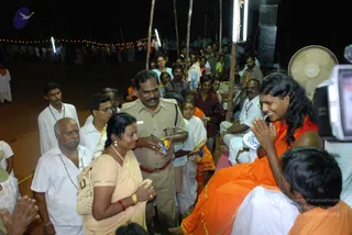 Night outdoor photograph of a public gathering with string lights and a standing fan at the edge. In the foreground, a long-haired person in orange and red robes sits on a raised mat with palms pressed together in namaste, smiling. Beside them, a uniformed police officer in khaki with a red-belted waist holds a small booklet, while a woman in a peach sari with gold jewellery and red bangles stands with hands folded. Men in white shirts and mundus, one with a garland and rudraksha mala, stand nearby, and a crowd in white and orange clothing stands behind. A camera operator is visible at the right edge.