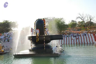 A photograph of a large black stone Shivalingam standing on a square platform in shallow green water, adorned with flower garlands in red, yellow and purple. Several men in white dhotis and white shirts, some with orange shawls, stand on the platform and in the water, pouring water and washing the lingam; one man in the water holds a long wooden pole. Water sprays up on the left. Behind, a crowd of white-clad people sits on striped red and white steps along the pool edge, with orange bunting strung across trees and a stone-lined embankment on the right.