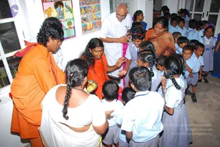 Indoor photo in a school corridor with white-painted walls and windows. A second saffron-robed figure with curly hair stands at left. A man in white shirt and white trousers with a cross-marked lanyard and a woman in an orange-brown sari stand behind the group. A woman in a white sari with a long plait faces away at lower left. Around them, many students in light blue-and-white school uniforms, some with ribbons and hair clips, crowd close, and a garland of pink flowers hangs from a child's neck. Educational wall charts are visible on the wall above.