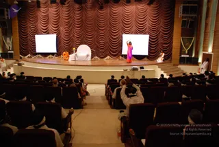 A photograph from the rear of an auditorium looking toward a raised stage with a brown swagged curtain backdrop. Two large blank projection screens flank the stage. On the stage, a figure in an orange robe sits beside a white chair with a small flower arrangement, and a woman in a pink top and orange skirt stands with one arm raised. A framed portrait with garland hangs at the stage side. Audience members in white clothing and white head coverings sit in rows of dark red seats, and one man in white raises his hands in front of him. Several people in white stand near the stage steps and at the right edge.
