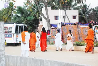 Outdoor scene on a dirt courtyard beside a white multi-storey building and palm trees. A white bus with a poster of a bearded man stands at left. Several people walk across the courtyard: a man in white dhoti and saffron shawl at left, a barefoot figure in saffron robes, a woman in a red sari with long dark hair, a man in white kurta and white dhoti, a woman in a white sari carrying a handbag, and a saffron-robed figure at right. Behind them colourful orange and red patterned cloth hangings and blue and pink bunting are strung on a stall. A printed poster of a woman in red hangs on a pole, and a small framed image is attached to the building.