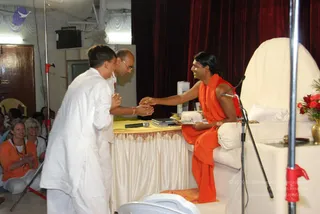A seated figure in an orange-saffron robe with long dark swept-back hair sits on a white-covered throne-like seat at a stage, smiling and extending his right hand to clasp the hands of a man in a white kurta standing before him. Beside them a second man in glasses and a white shirt watches at a table draped in yellow cloth with a microphone, notebooks and a red flower arrangement. Women in orange and white sit on the left, and a red curtain backs the stage.