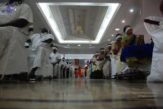 A photograph taken from floor level inside a large hall with a recessed ceiling, a central chandelier and bright ceiling lights. Rows of seated people in white uniform-style clothing with white headbands or eye-covers line both sides of a central aisle, many with red-trimmed white cloths. On the right, women in saris, including one in blue and gold and one in yellow, sit with white blindfolds. A standing figure in orange robes stands at the far end of the aisle near a banner with a portrait. Tripod equipment rests on the floor at right.