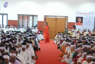 A large indoor hall with white walls, barred windows and a wooden door. Hundreds of seated participants wear white blindfolds over their eyes, most in white or pale clothing with some in saffron, orange or pink saris. A standing figure in a long saffron-orange robe walks down a red carpet between the rows, with a clear view of the row of seated attendees on both sides. The scene is a structured group practice in a meeting hall.