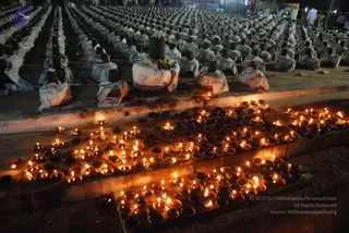A night photograph of a large outdoor ritual field filled with rows of black Shiva lingams, each draped in white cloth with coloured borders, many topped with flower garlands. In the foreground, hundreds of small clay oil lamps with flickering flames line the stone steps and ground. Standing devotees are visible in the far background beneath festive lights, and the scene reads as a mass abhishekam or lingam puja arrangement. No identifiable person is shown in the foreground.