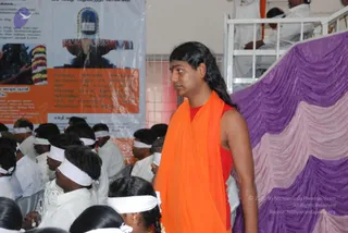 A photograph inside a hall where a standing figure in an orange shawl over a red sleeveless top, with long swept-back dark hair, stands in front of a purple and pink draped stage. Behind him seated devotees in white clothing wear white headbands across their foreheads. A white railing and a poster with a Shiva lingam image and Tamil text appear at left, and a man in white stands at the upper right. The standing figure shows distinctive long hair and orange saffron clothing, so he is a probable Swamiji in person.