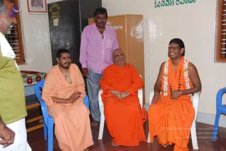 A photograph of four men in a small room with cream walls and a tiled floor. On the right, a smiling man in saffron-orange robes with a white flower garland and a rudraksha mala sits on a white plastic chair; his hair is swept back and he has ornate gold-toned adornments. Beside him, a bald, mustached man in orange robes sits with hands folded. On the left, a seated man in peach-orange robes and a pendant necklace looks to the side, and a standing man in a pink shirt and grey trousers with a gold chain stands behind. A man in green and white is partly visible at the left edge. Blue plastic chairs and a framed picture on the wall are visible, along with Kannada and Om Namah Shivaya lettering. Probable Swamiji is the smiling man on the right, identified by his swept-back hair, saffron robes, rudraksha mala and garland.