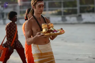 A shirtless young man with dreadlocks and a white tilak on his forehead carries a gold tray with two ornate gold vessels, flowers and red offerings across a pale open courtyard. He wears a white dhoti with gold borders, a red bindi and a rudraksha mala with a pendant. Behind him, a woman in an orange sari with a red hair ribbon walks, and to the left a person in a maroon patterned kurta carries an orange bag. Late sunlight warms the scene.