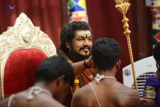 A seated man in saffron-orange robes with rudraksha malas and a gold necklace sits on an ornate gilded throne with a red velvet seat, carved with a face motif. He wears a white tripundra with a red bindi on his forehead and smiles while gesturing toward two bare-backed young men seen from behind, one with a white sacred cord over the shoulder. A tall gold ceremonial staff stands at right and a red and white garland hangs near his chest. Red curtains and colourful banners form the backdrop, with a white fan at right and a woman in purple at the edge of the frame. This appears to be a ritual or darshan setting. The seated man is an appearance-based a person candidate, identified by his saffron robes, rudraksha malas and gold necklace while seated on the throne.