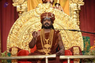 A seated man in a rust-brown saffron-toned robe speaks into a black microphone on a stand. He wears a rust-orange turban with a gold ornament at the front, a gold-toned multi-strand necklace with a large pendant, gold bracelets and armlets, and a white-and-ash forehead mark. He sits on an ornate gold-leaf throne with red cushions and armrests, his right hand raised in a gesturing mudra. Behind him, gold-and-red drapes, brass lamps and two figures with crowns appear partly visible. Orange flowers and greenery line the lower edge.