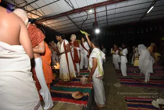 Night photo of a homa in an open-sided shelter with a corrugated metal roof and string lights overhead. A small fire burns at left beside the fire pit, with brass and clay vessels nearby. In the foreground a shaven-headed man in a white dhoti stands with his back to the camera. Beside him, a man in an orange-saffron wrap stands near the fire. A woman in a white sari with a red border and gold trim holds a white and gold garland, and a blonde woman in a red shawl and white outfit stands with palms joined in prayer. A young man in white dhoti and shawl raises a green leaf or ritual item. Other participants in white clothes sit or stand on striped red, green and blue mats across the floor. A darker wrap and white ritual garments fill the background, with the mats and floor marked with kolam patterns.