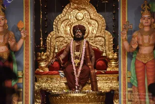 A seated figure in an ornate gold throne (peetha) with a gold-embossed back and a red cushion, wearing a rust-brown turban with a tilak, rust-brown robes, a draped brown shawl, a pink-and-white flower garland, and a gold necklace with a pendant. Eyes closed, hands resting on the knees. Flanking the throne are two painted deity murti figures with multiple arms, crowns and gold jewellery. Brass oil lamps (deepa) and bells hang at the sides, and a small offering tray with flowers and plates sits at the base.
