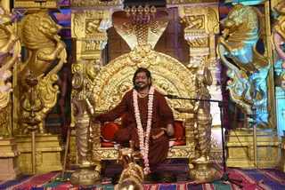A man sits cross-legged on an ornate golden throne with a cobra-hood canopy and carved lion-shaped armrests, smiling at a microphone on a stand. He wears a rust-red robe and a long white and pink flower garland. Flanking him are golden lion sculptures, lamp stands and ornate golden pillars, with a colourful striped cloth on the floor. Swamiji (probable): the figure has long dark hair and a rust-red robe with a flower garland, seated on a golden throne at a discourse stage. A golden deity figure stands at the lower edge of the frame.