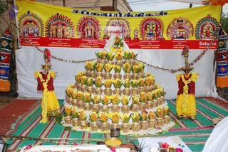 A photograph of a ritual display at an outdoor venue. A large pyramid of tiered brass pots, each wrapped in woven netting and topped with yellow flowers and green leaves, is arranged on a white cloth atop a striped mat. Two standing brass lamps flank the arrangement, one on each side, each garlanded with yellow and pink flowers. Behind it hangs a yellow and red banner with painted panels of deities in arched frames, labelled with Tamil and English names and chakra designations. Brass vessels, flowers and a bowl of yellow liquid sit in the foreground. No people are visible in the frame.