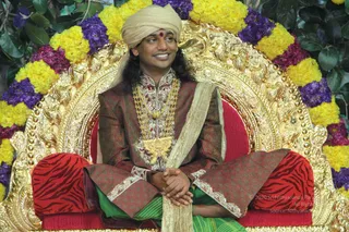 A photograph of a seated man on an ornate gold throne framed by garlands of yellow marigolds and purple flowers with green leaves behind. He wears a cream turban with a red bindi, a maroon and gold embroidered silk kurta with a heavy gold necklace and pendant, and a cream shawl draped across his body. He sits cross-legged on red cushions with zebra-stripe patterns, hands clasped in his lap, smiling and looking to the side. a person by appearance cues of ornate ceremonial gold necklace, turban and elaborate silk attire seated on a throne; the throne alone does not confirm identity.