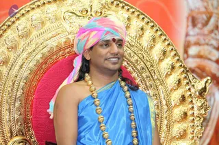 A seated man in a multicoloured pink, blue and turquoise turban and a blue silk shawl with a gold border sits on an ornate gold throne with a red cushion. He wears a heavy ceremonial necklace of gold beads and a red tilak on his forehead, and smiles toward his right. Behind the throne is a blurred orange and red backdrop. Swamiji (probable): the appearance cues are the colourful turban, ceremonial gold beaded necklace and blue shawl on a gold peetha, and the page names him as the speaker.
