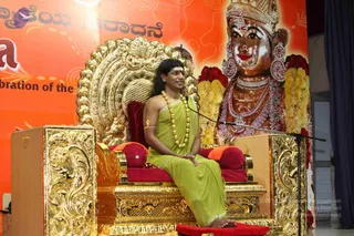 A man with long dark hair, a red tilak on his forehead, a gold rudraksha-style mala and gold bangle sits cross-legged on an ornate gilded throne with red cushions, wearing a light green garment draped over his shoulder. He sits before a microphone stand at a satsang. Behind him a large backdrop shows a golden deity murti of a goddess with a crown, red garland of flowers, and jewellery against an orange background. a person appears on the throne, with long swept hair, mala and green ornate draping; he is a speaker at the event.