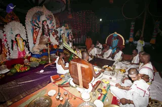 A night-time puja at an outdoor pandal lit by string lights. Behind a worship area stands a large decorated deity murti with multiple raised arms, a lion beside it, and smaller white-framed deity figures with garlands. Offerings of fruit, a plate of watermelon and coconuts, and brass lamps sit on a purple cloth. Several men in plain white dhotis and gold-bead necklaces sit cross-legged around a low wooden yantra-like platform with a fire pit and bowls. A man in a white vest and bare chest sits near a wooden drum-like board. A figure in a blue garment and turban sits on a gold-and-red throne at the back right, and a priest in orange robes stands behind the group. Several others in white attend the scene, and red-lit decorations and chariot-style posts frame the background.