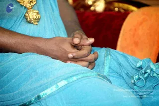 Close photo of a seated person's lap and hands, clasped together in front of a turquoise-blue garment with a shimmering teal border. A large gold ornamental pendant hangs at the upper left over the blue fabric. Behind, a red cushion and an orange cushion fill the background. Only hands, forearms and clothing are visible; no face is shown. The clasped-hands posture suggests a seated satsang setting, though no identity can be assigned from appearance.