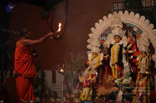 A photograph of a temple-style shrine at night. A man in saffron-orange robes with a shaved head stands at left, extending his arm to hold a lit oil lamp toward a deity murti. The central goddess figure has a white crown with red and gold accents, multiple arms holding weapons, and is garlanded with yellow marigold and white garlands. She stands beside a lion. Flanking figures wear white and red garlands, a small elephant-headed figure appears at lower centre, and brass oil lamps stand on the altar with a potted plant and banners behind.