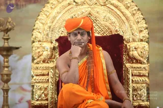 A seated man in saffron-orange attire sits on an ornate gold throne with carved lion figures and a deep red velvet cushion. He wears an orange turban with gold trim, an orange shawl with gold border, a red-beige tilak on his forehead, a beaded rudraksha-style mala and gold bangles, and a small headset microphone. His right hand rests against his cheek in a contemplative pose. Behind him stands a gold ornamental lamp stand. Appearance-based probable Swamiji: the orange turban, saffron shawl, beaded mala and throne are visible cues of a seated spiritual teacher during a discourse setting.