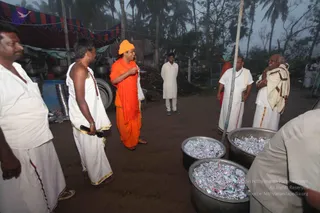 Outdoor relief-camp scene on bare ground beneath palm trees and a striped tent canopy. A man in an orange turban, orange upper cloth and orange dhoti stands centre, gesturing with one hand and smiling. Around him, several men in white dhotis and white shirts or shawls stand talking; one holds a rudraksha mala. Two large steel pots in the foreground are filled with foil-wrapped packets, and cooking vessels and drums are visible behind. Men in white are attendants or devotees.