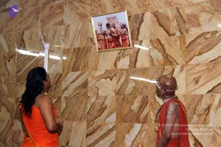 A photograph of two people in orange-saffron robes standing before a marble wall with a framed photo of four sadhus in orange robes in front of a temple gopuram. On the left, a woman with long black hair wears an orange sari and gold jewellery, facing the framed picture. On the right, a bald man in an orange shawl with a rudraksha mala and a gold necklace looks up at the framed photo. The marble wall has warm brown veining, and the framed portrait hangs slightly above eye level.