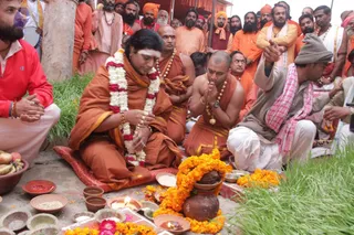 Outdoor ritual scene on grass. A seated man in orange robes with a white-and-red garland and a white forehead tilak sits on a red cushion with eyes closed and hands folded in prayer, while a shaved-headed man in orange robes and gold beads beside him also presses palms together. Around them stand many men in orange, saffron and white clothing, some wearing orange turbans and rudraksha malas. In front lie brass and clay bowls with red powder, grains, flowers, bananas, a clay pot wrapped in marigold garlands, and oil lamps. A man in a grey cap and pink-checked shawl sits at right with hands joined. Identification of the central seated figure is uncertain from appearance alone.