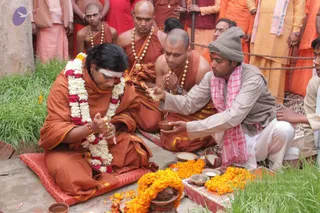 Outdoor puja on grass beside a stone platform scattered with marigold flowers. A seated figure in orange robes and a white-and-red flower garland, with a white tilak on the forehead and a long dark hair, sits on a red cushion with hands joined in a mudra. Beside him, a man in a grey knitted cap, grey vest and pink-checked cloth holds a small brass plate and offers items. Behind them, monks in orange and saffron robes with rudraksha malas stand and sit, and a crowd in orange and yellow stands behind a rope barrier. Marigold heaps, small bowls and a dark stone object lie on the platform. a person: the seated figure wears a long-haired, orange-robed appearance with a garland and tilak, consistent with the page's reference to a person.