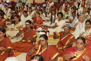A seated group of devotees performs a ritual with lit oil lamps in a hall. Front left, a woman in an orange-red sari with gold border and rudraksha mala holds a lamp, and a woman in orange with white forehead markings lifts a small flame near the centre. Others in white and red saris hold small lamps and brass vessels on orange mats. Behind them, rows of participants in white and orange clothing sit on the floor and red chairs, many with rudraksha malas and tilak markings. The scene suggests an aarti-style lamp offering in a satsang setting.