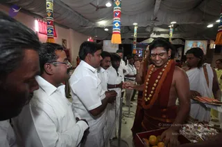 A photograph inside a decorated hall with a white tent-style ceiling, hanging colourful striped pillars with tassels, and framed religious pictures on the walls. A man in a red-orange robe with an orange shawl, rudraksha-style gold bead necklaces, a white tilak on his forehead and a shaved head stands at right, smiling and handing something to a devotee. A fruit basket with oranges sits at lower right. Devotees in white shirts and white dhotis line up on the left, some with hands folded. A man in a white dhoti and shawl stands at far right holding a tray.
