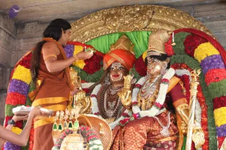 A figure in a rust-orange dhoti and matching shawl with a gold-coloured sacred cord and gold armlets stands at left, long dark hair falling down the back, offering flowers from one hand over two golden murtis seated on a throne. The seated golden figures wear crowns or mitres, gold necklaces and jewelled garlands; the left figure has an orange turban and the right wears a red-and-gold brocade garment and holds a golden staff. The throne is framed by a golden arch, a green backdrop and dense bands of red, yellow, white and purple flower garlands. At bottom left a hand holds a silver tray of offerings. The scene resembles a puja or abhishekam. a person is not clearly identifiable, as the standing figure's face is partly turned and the appearance is uncertain.