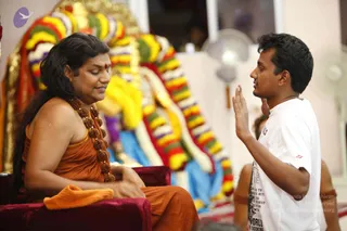 A seated figure in an orange-saffron garment with a long, loose-flowing dark hairstyle and a large rudraksha mala with a gold-toned ornament sits on a deep maroon cushioned seat, with a small red bindi on the forehead. A young man in a white t-shirt with dark printed graphics stands facing them, one hand raised in a gesture as he speaks. Behind them is a large decorative backdrop of garlands in yellow, red, green, blue and white, with a golden crown-like top and a pedestal fan on the right. An orange cloth rests on the seat beside the seated figure. The appearance-based cues of long hair, saffron clothing and rudraksha malas are consistent with a person seated in discourse, though this is uncertain.