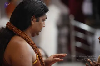 A close side profile of a man with long dark hair swept back, wearing a thick rudraksha mala of brown beads around his neck and an orange-brown garment with a yellow edge at the shoulder. He is gesturing with an open hand toward someone just out of frame at the right edge. The background is blurred with indistinct figures and a pale wall.